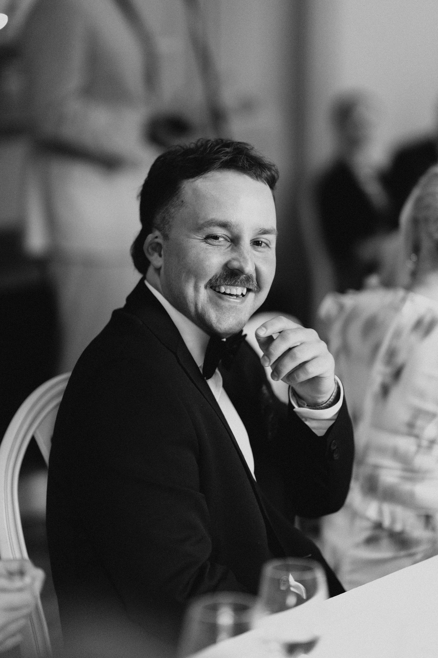 Smiling man in tuxedo with bow tie enjoying wedding reception dinner, black and white portrait.