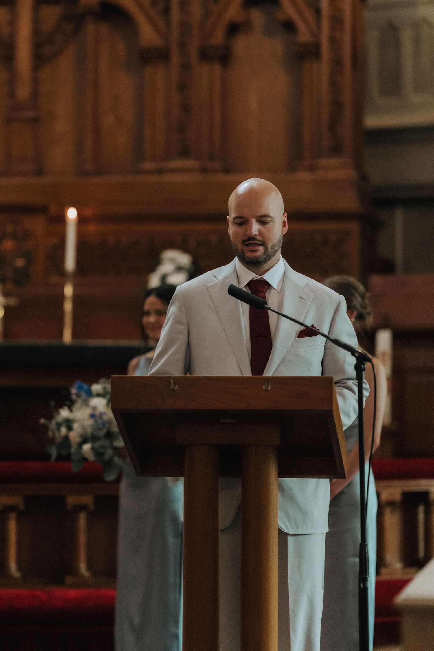 Bald man in white suit with burgundy tie delivering a reading at a wooden church pulpit during a wedding ceremony.