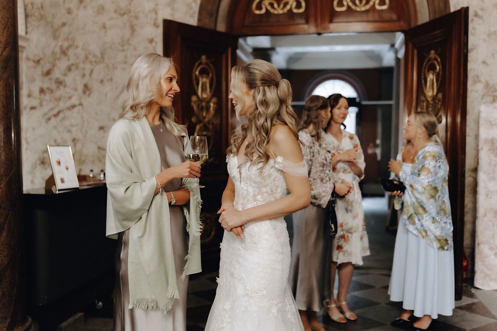 Bride in white lace gown greeting guests near ornate wooden doors at wedding reception.