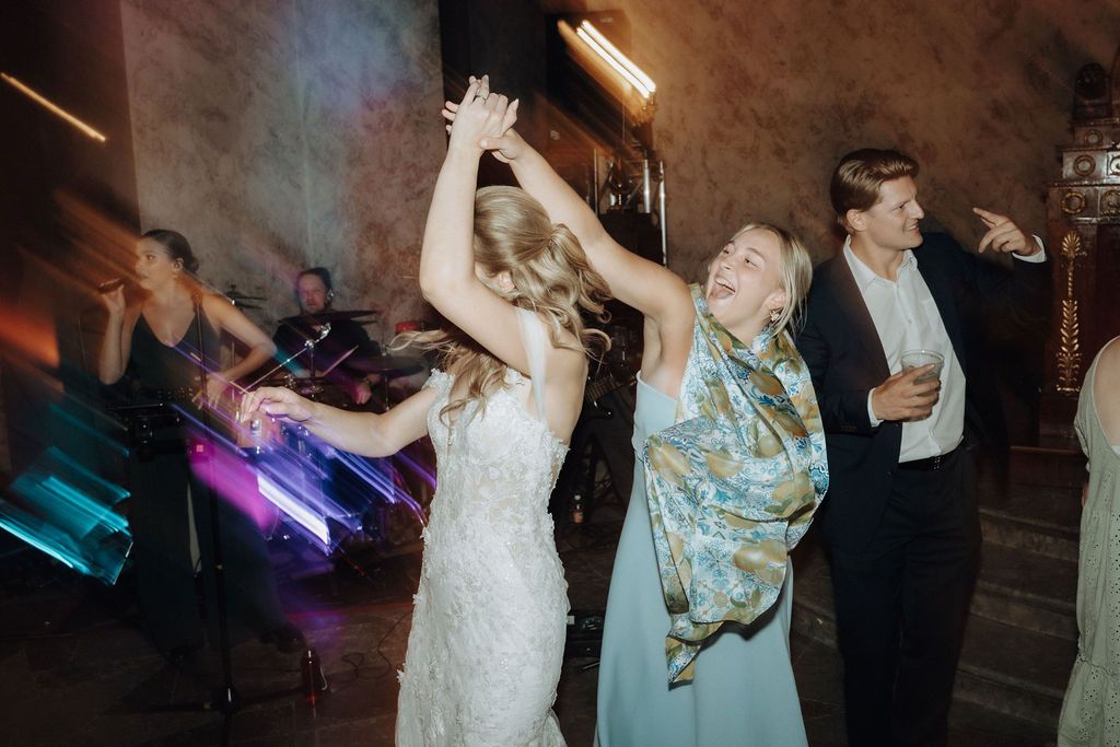 Bride dancing joyfully with female guests at a wedding reception with live band performing.