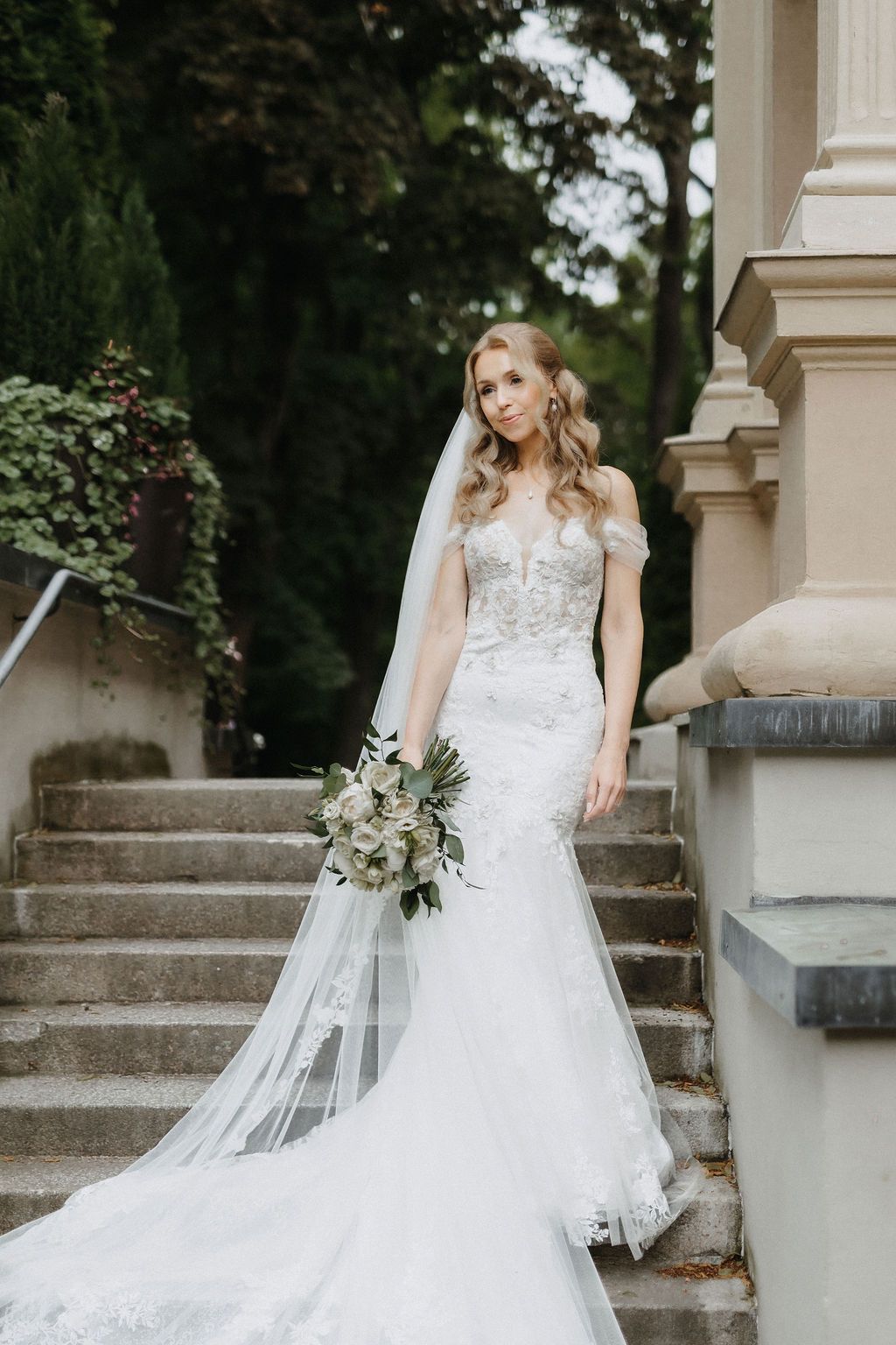 Elegant bride in fitted lace mermaid gown holds white bouquet on outdoor stone staircase.
