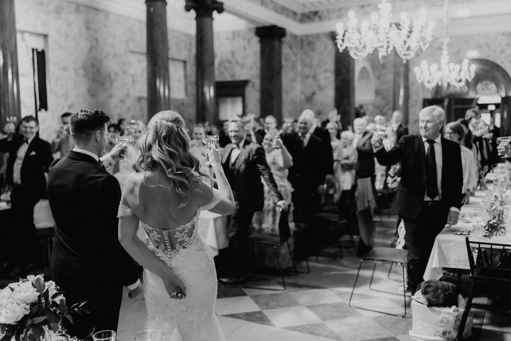 Black and white photo of bride and groom facing guests in grand chandelier-lit ballroom.