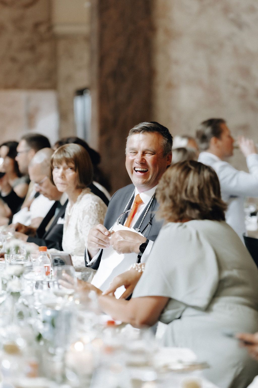 Laughing wedding guest in gray suit with orange tie enjoying reception dinner at long table.