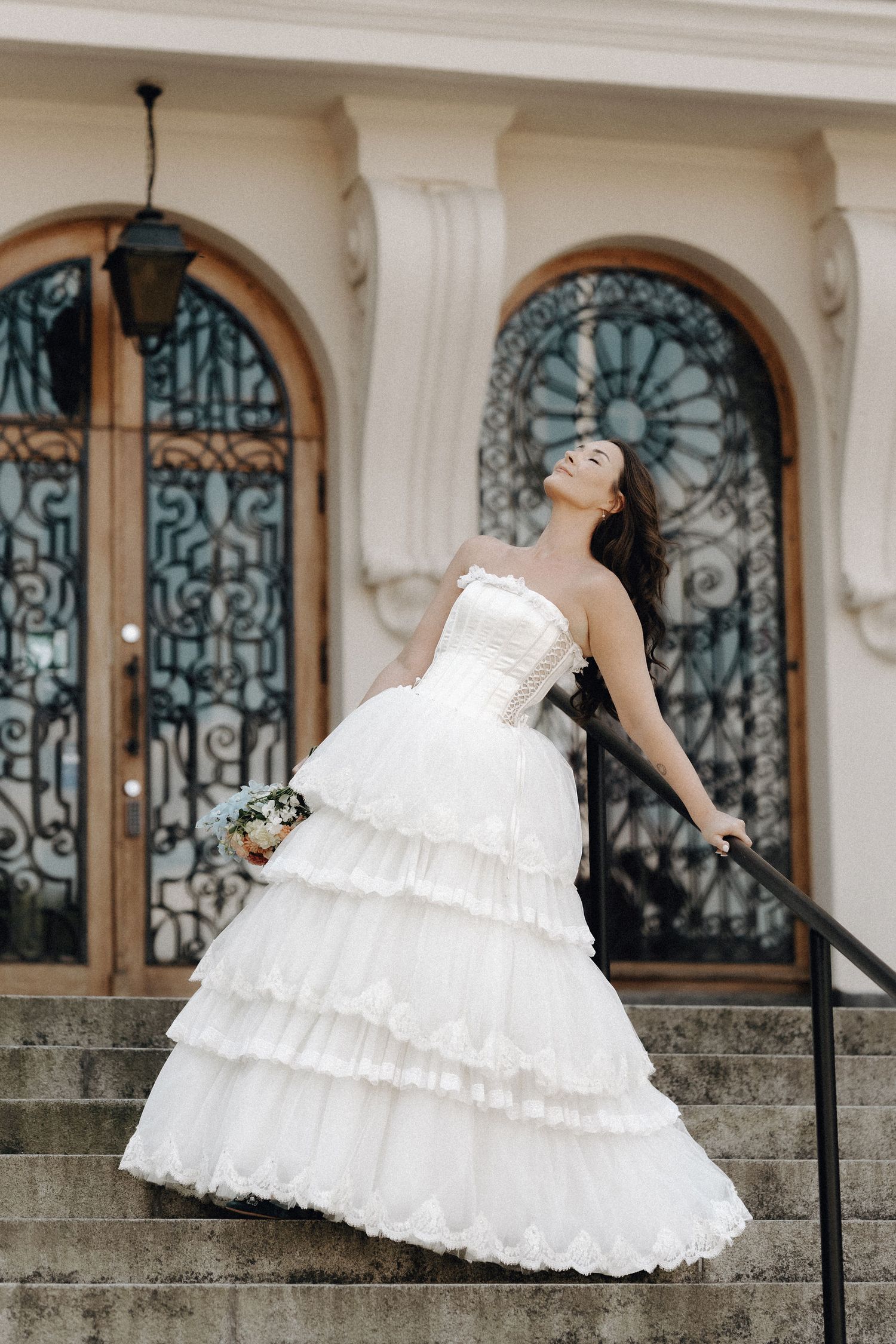 Bride in a ruffled white ball gown posing gracefully on stone steps outside an ornate building.