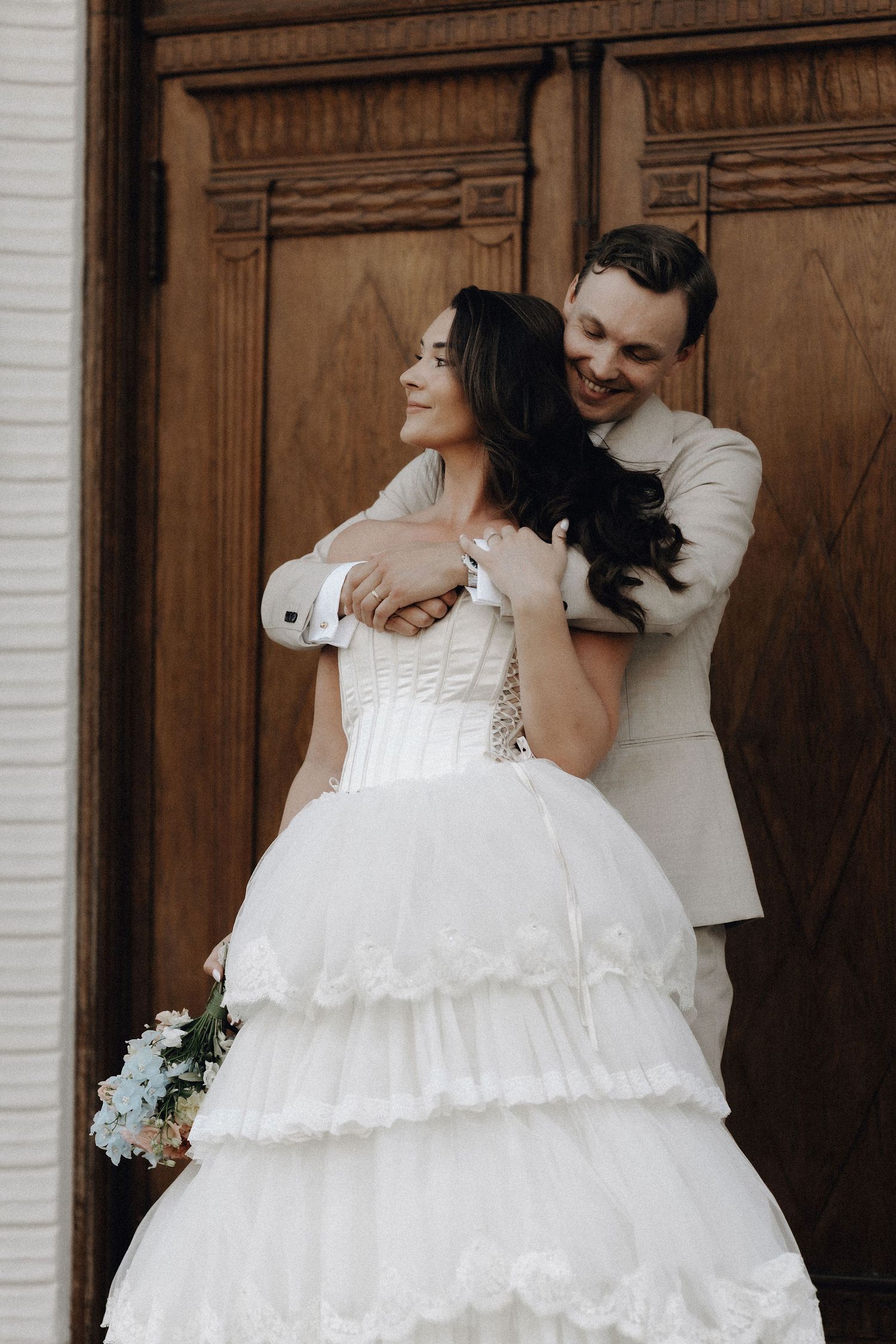 Groom embracing bride in layered white wedding gown before elegant wooden doors.