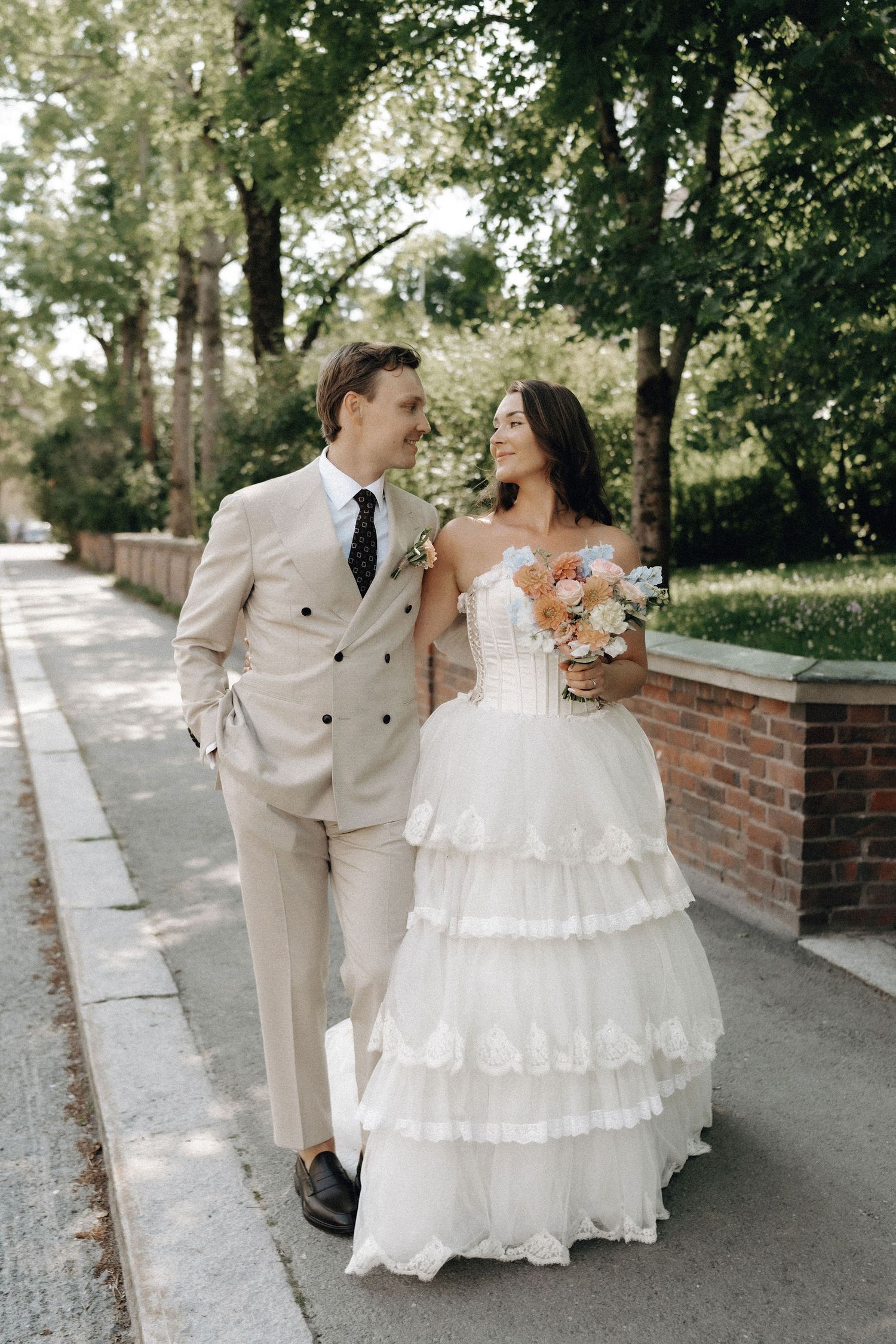 Smiling newlyweds walk closely together on a tree-lined sidewalk, bride carrying a pastel bouquet.