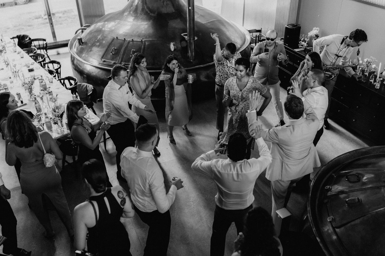 Aerial black and white view of wedding guests dancing around a brewery tank.