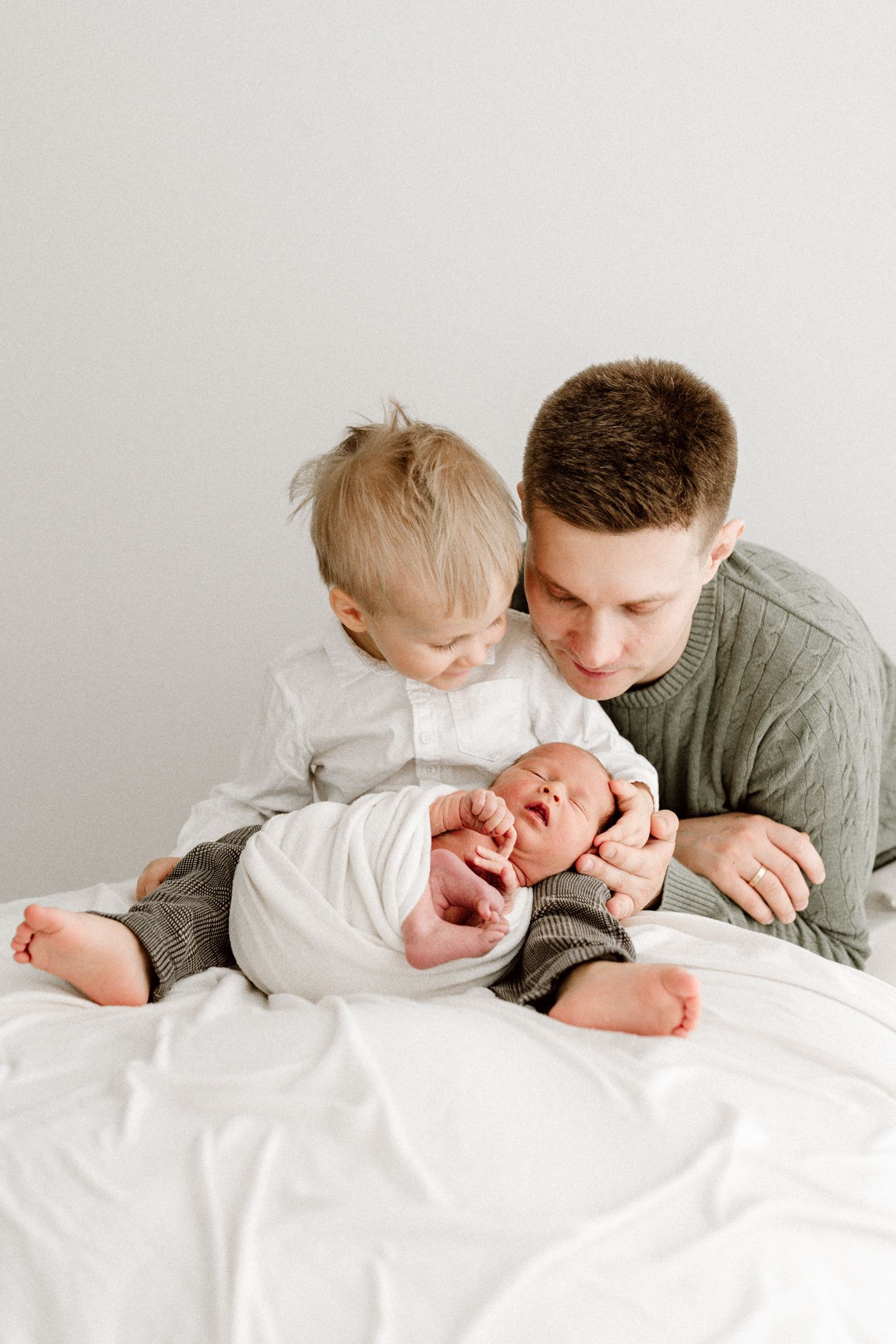 A father in a green sweater and his toddler son sit together on a bed with their newborn baby.