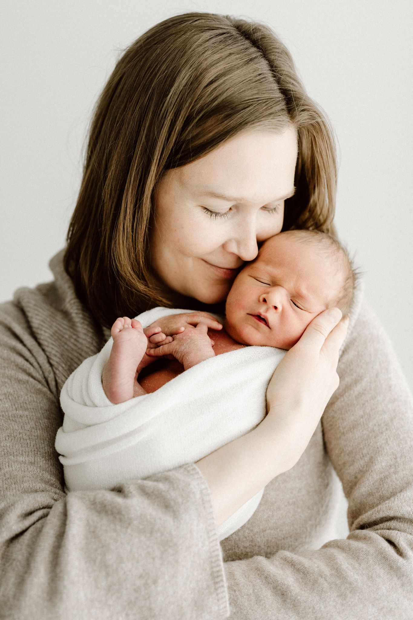 Loving mother in beige sweater gently pressing her cheek against her sleeping swaddled newborn baby.
