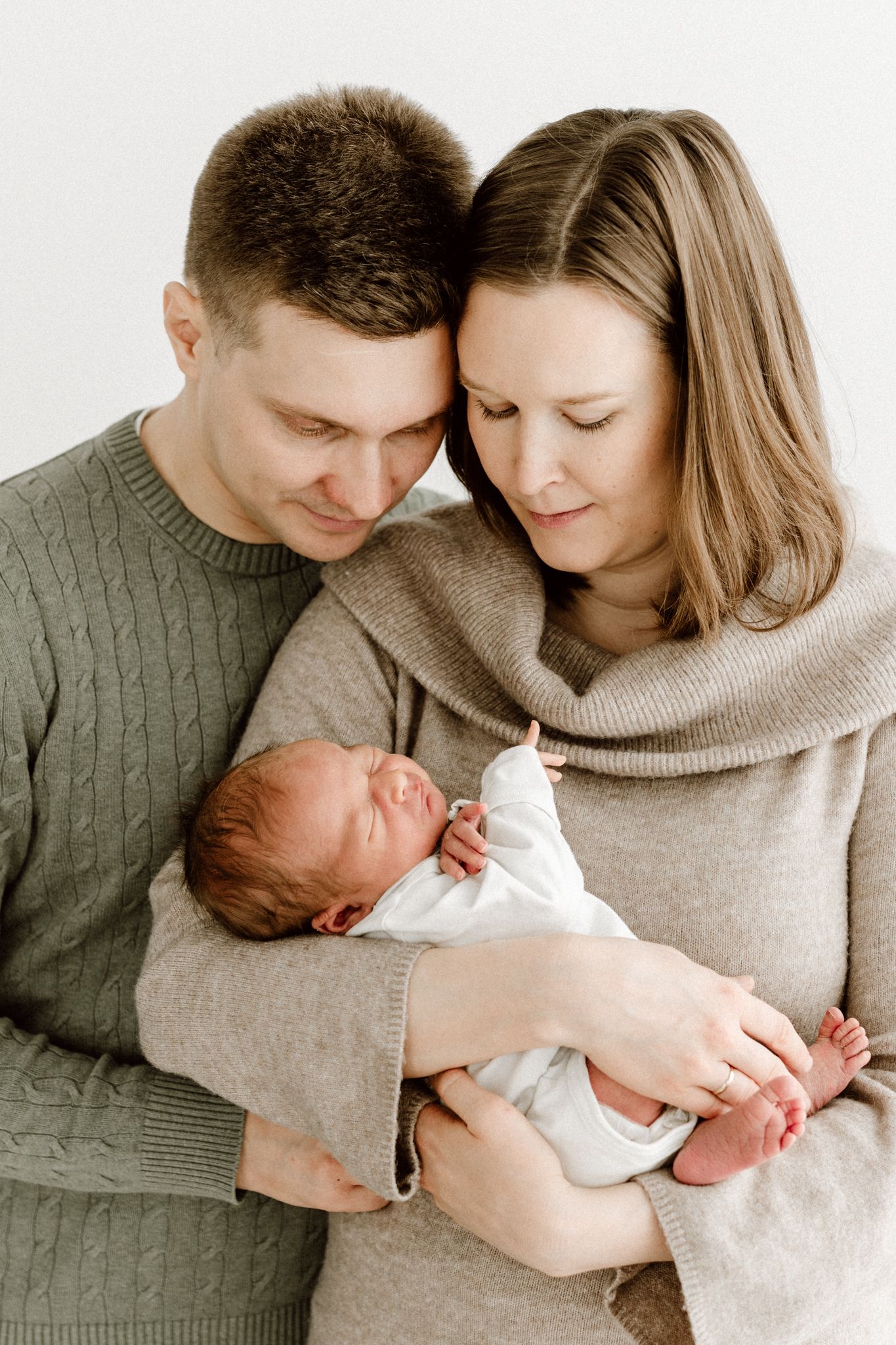 Happy parents in neutral tones holding their sleeping newborn baby close together against white background.