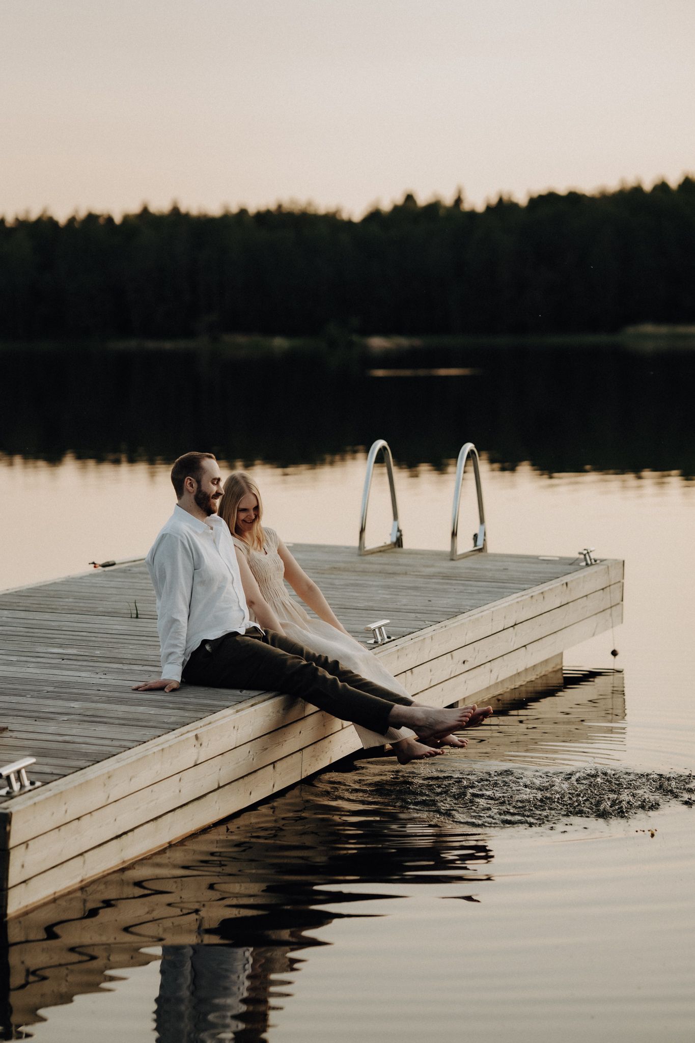 Couple sitting on a wooden dock at sunset, feet dangling over a calm lake with forest backdrop.