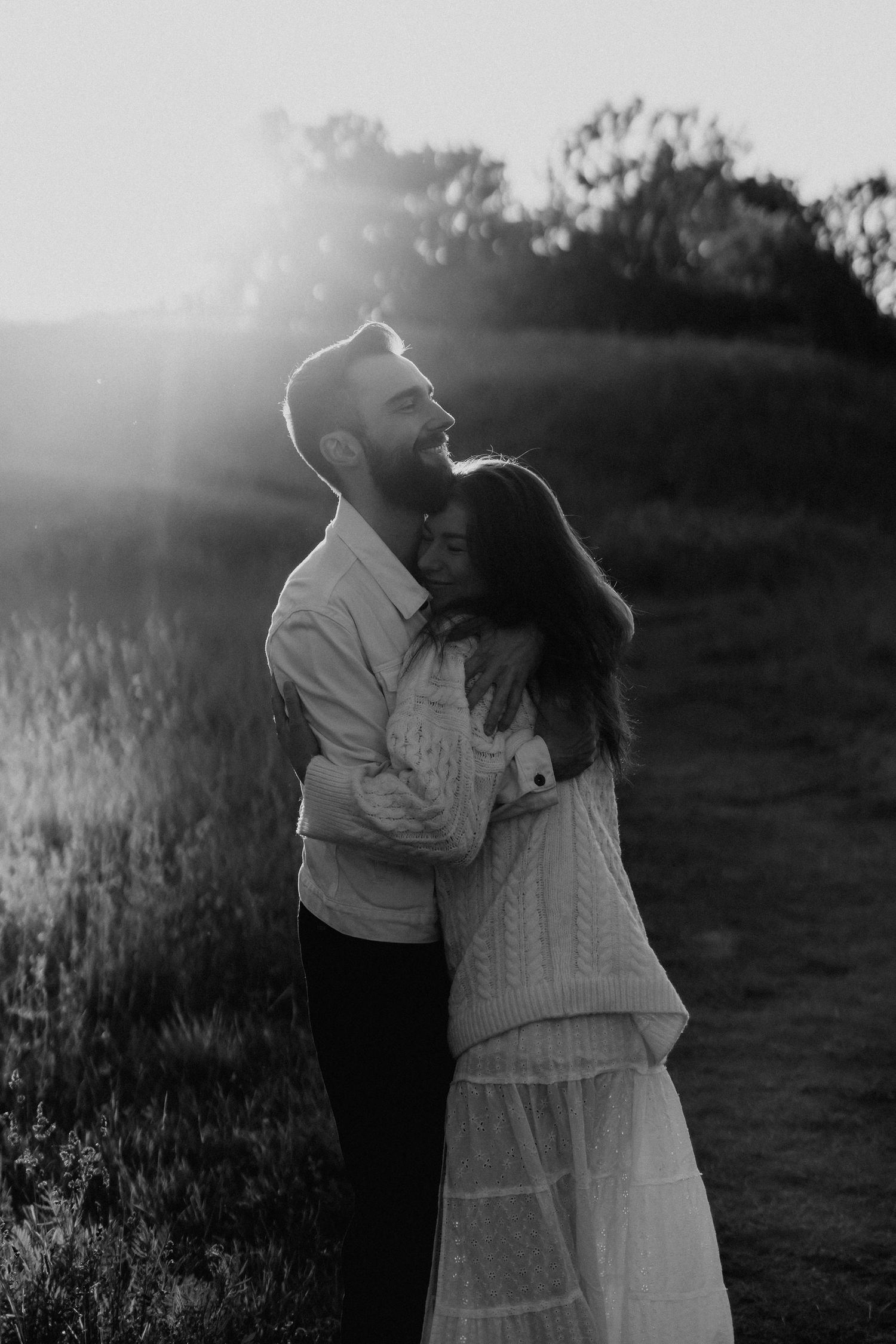 Black and white photo of a couple hugging outdoors at sunset, man laughing joyfully.
