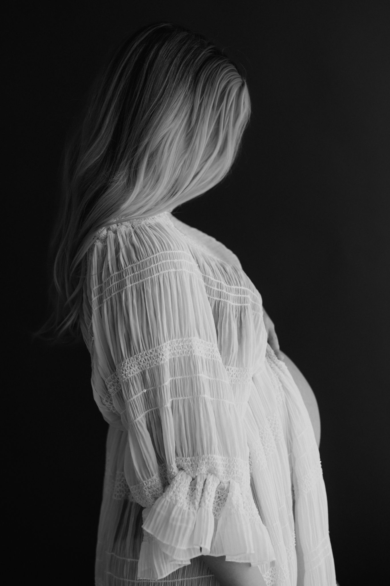 Pregnant woman with long blonde hair in white lace dress photographed from behind, black and white maternity portrait.