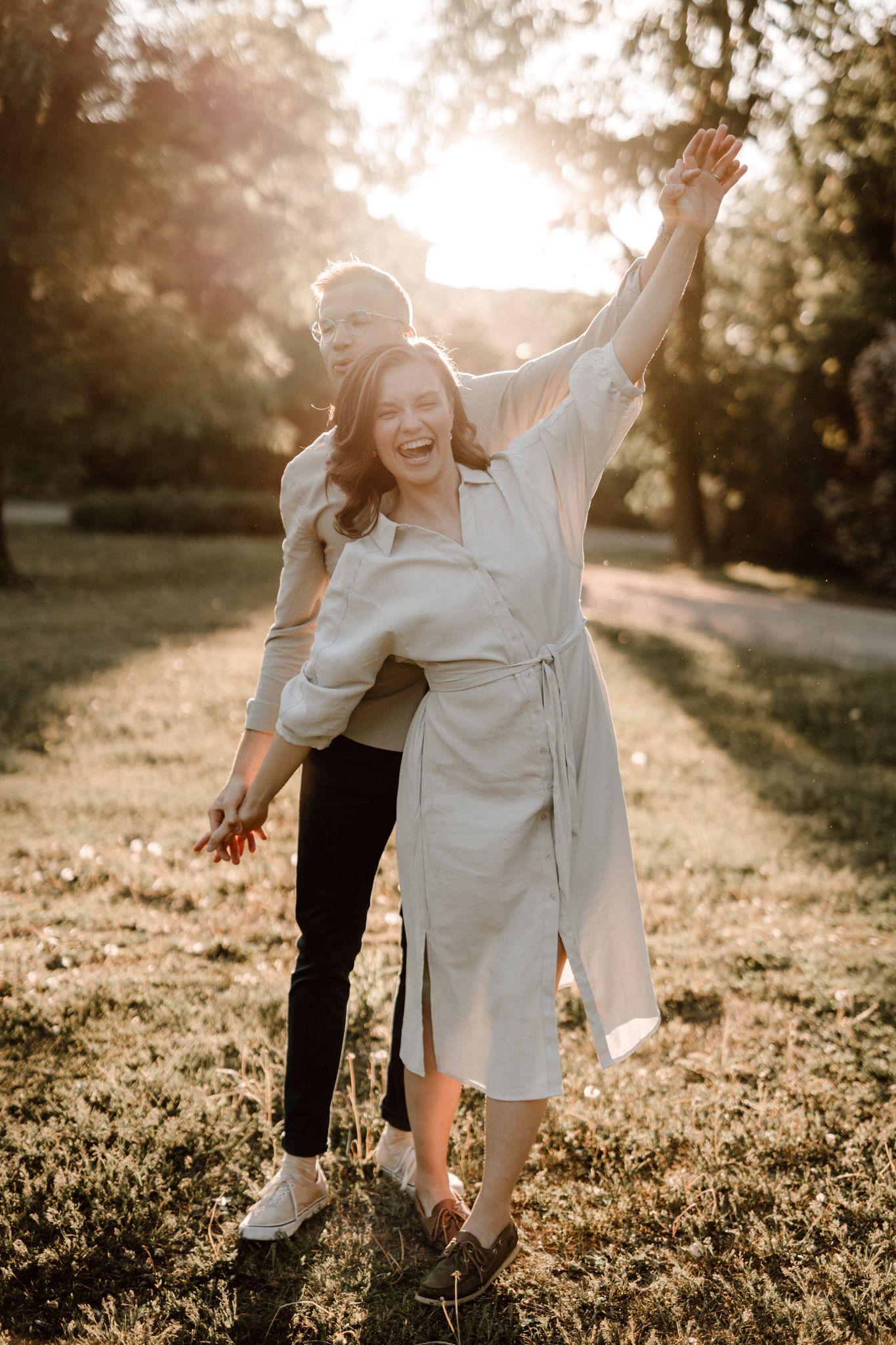 Laughing couple playing in a sunlit park, woman raising her arm joyfully as man hugs her.