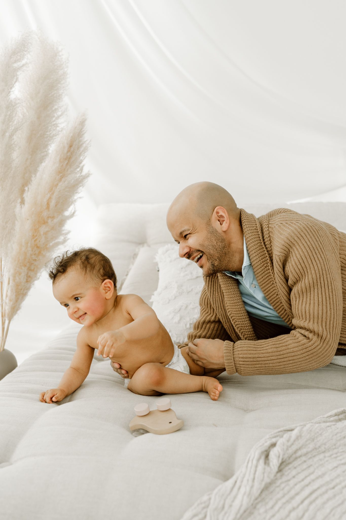Smiling bald father in brown sweater bonding with baby on white quilted bed.