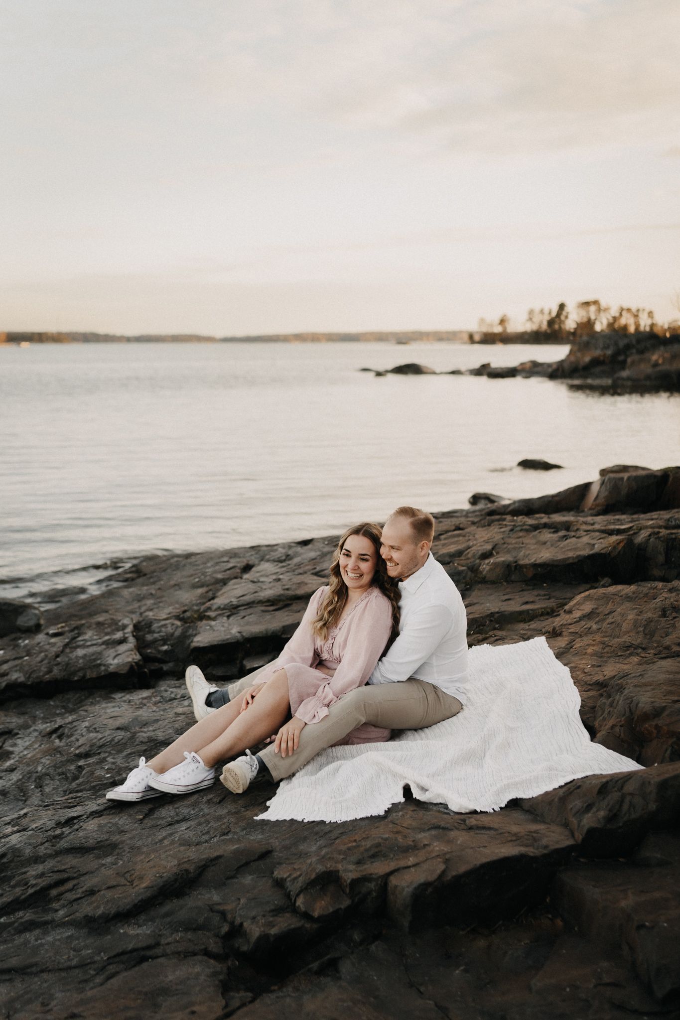 Happy couple sitting together on white blanket over rocks by a serene lake at golden hour.