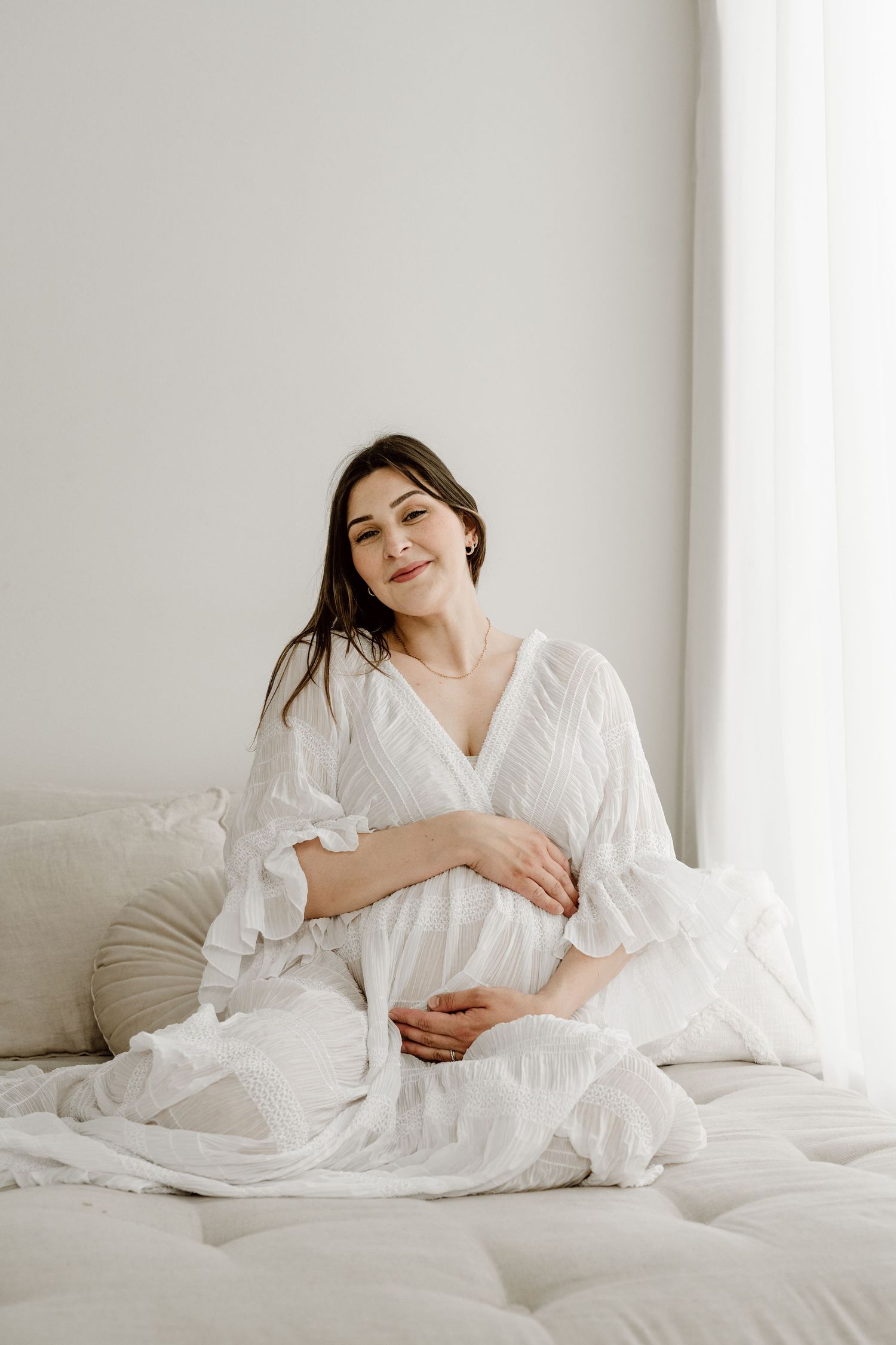 A serene pregnant woman in a white dress sits on a bed cradling her belly near a bright window.