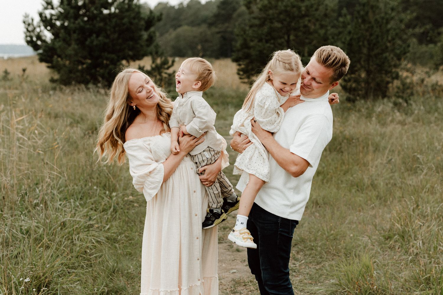 Smiling family of four holding two young children in a sunny meadow outdoors.