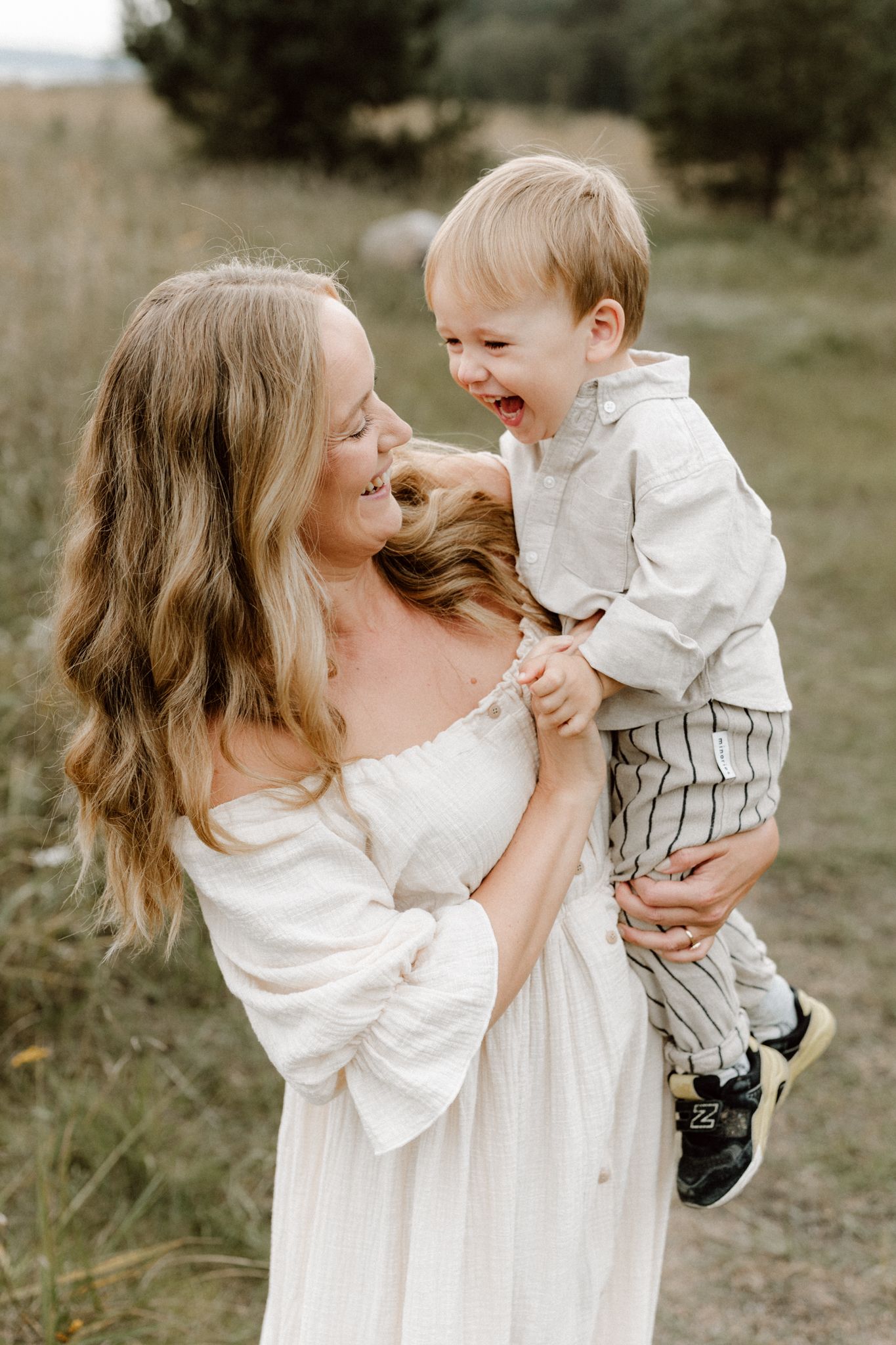 Smiling blonde mother in white dress holding laughing toddler boy in field.
