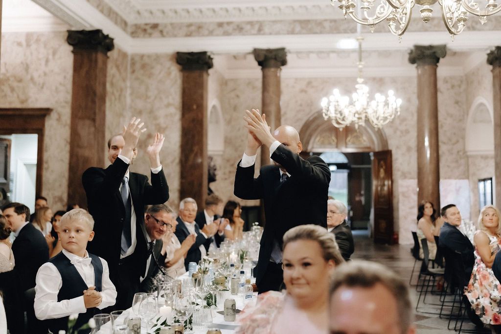 Wedding guests raising hands and clapping in a stunning chandelier-lit marble reception hall.