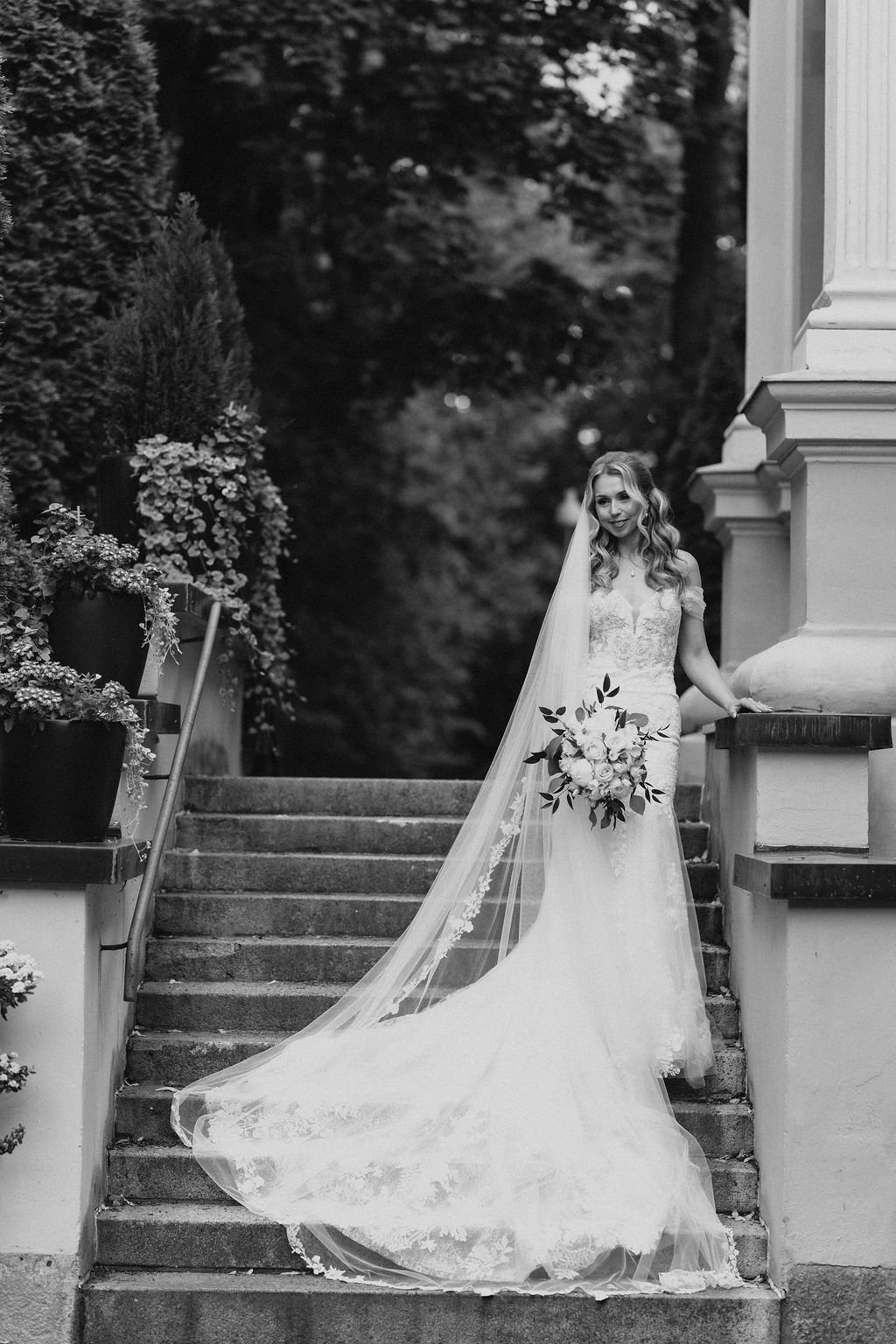 Bride in lace gown and long veil poses on stone steps surrounded by lush greenery, black and white photo.