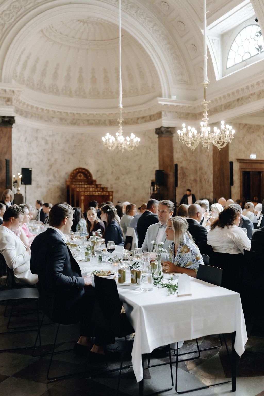 Wedding guests seated at white linen tables in ornate domed reception hall with chandeliers.