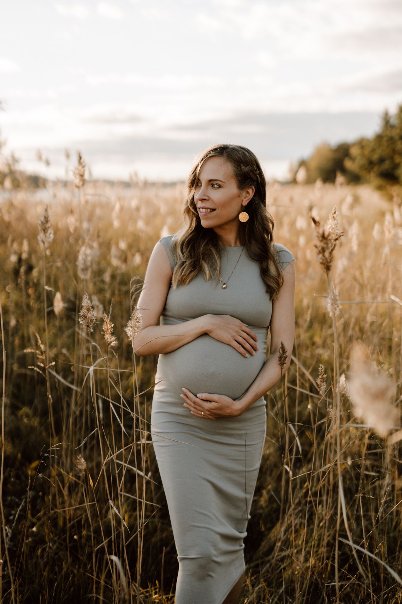Smiling pregnant woman in sage green dress cradling belly while standing in tall golden grass field.