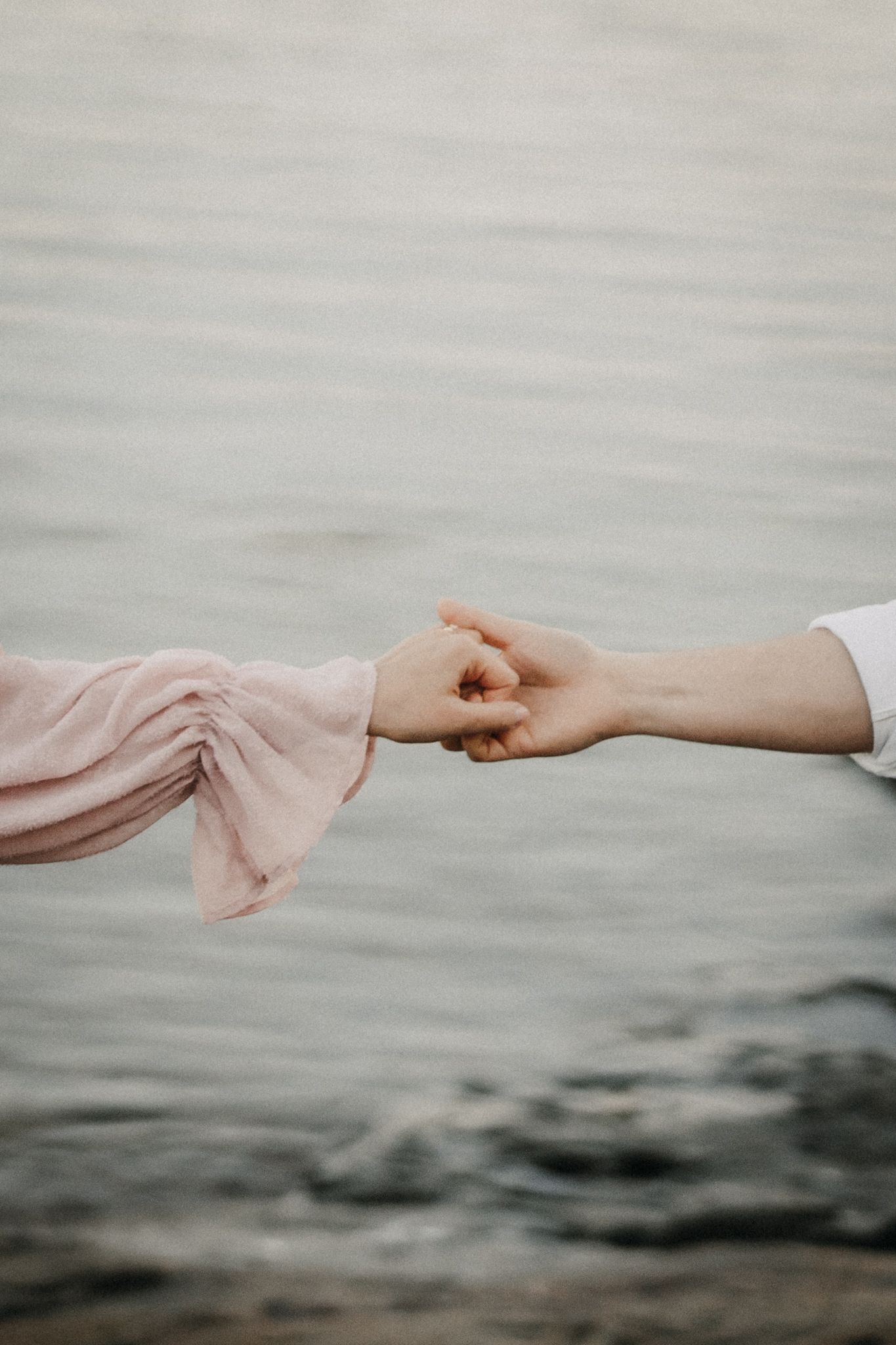 Close-up of couple gently holding fingertips over calm water, woman in flowing pink sleeve dress.