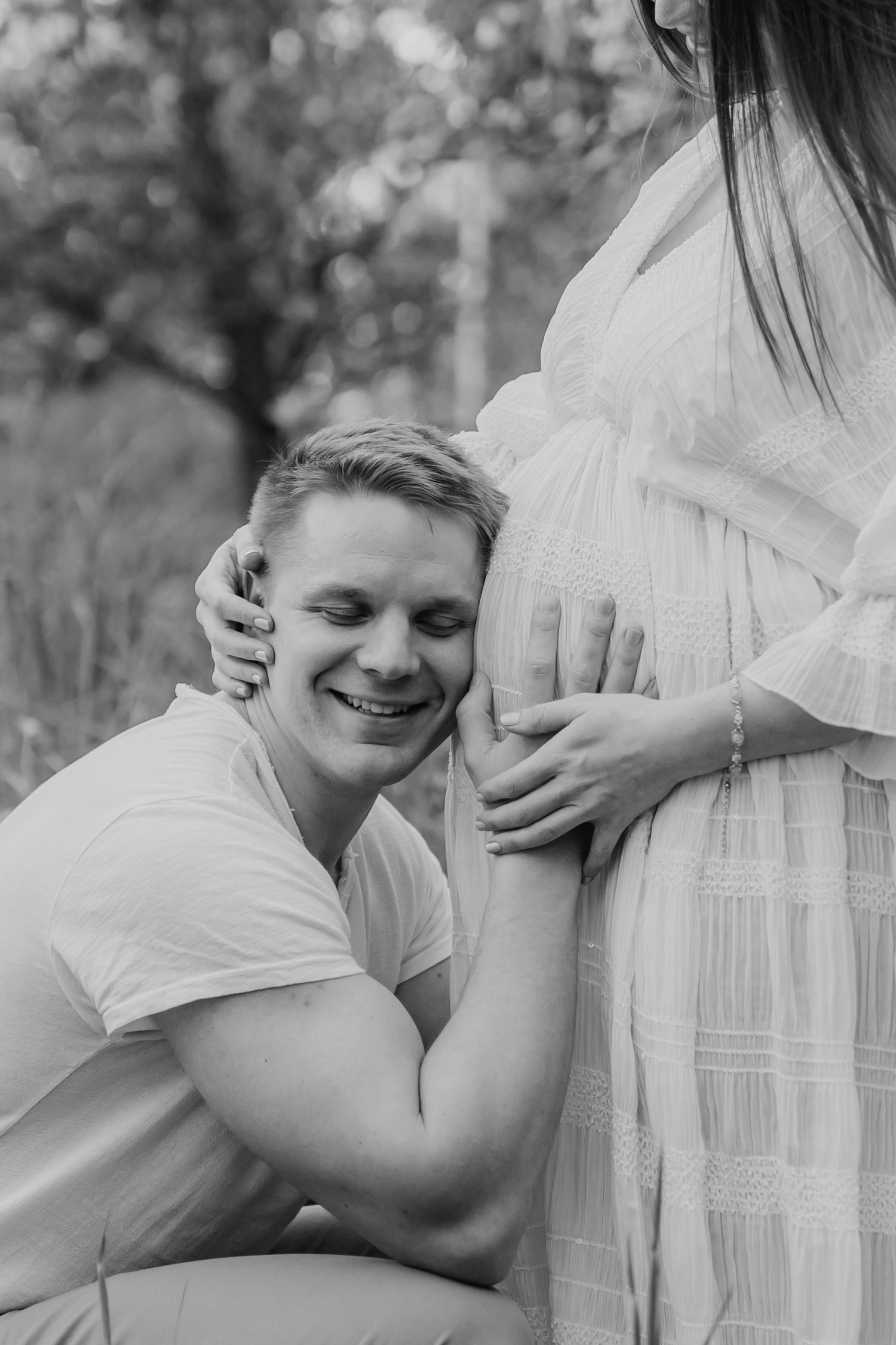 Black and white photo of smiling man kneeling to listen to pregnant partners belly in outdoor setting.