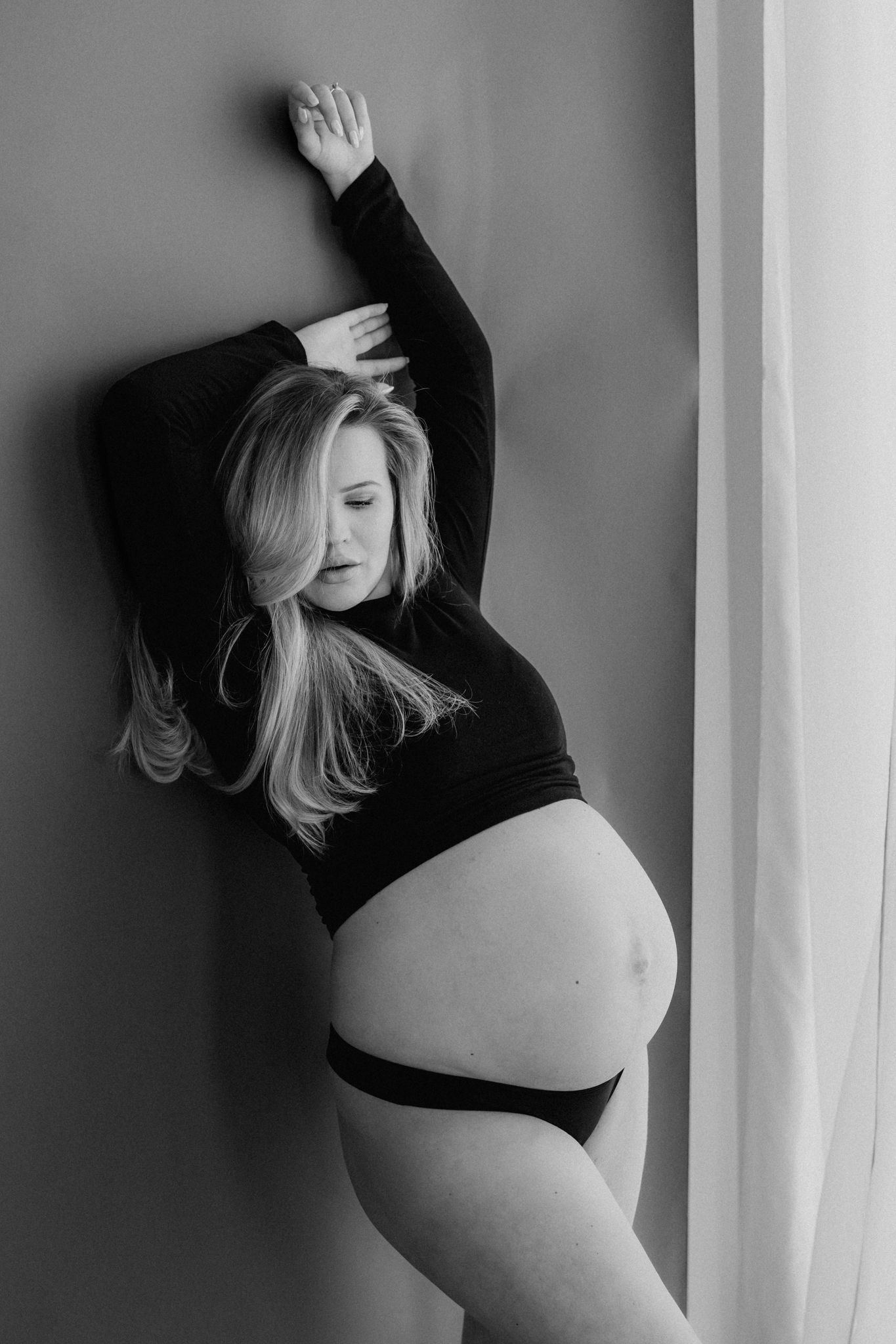Black and white maternity portrait of pregnant blonde woman leaning against wall with arm raised.