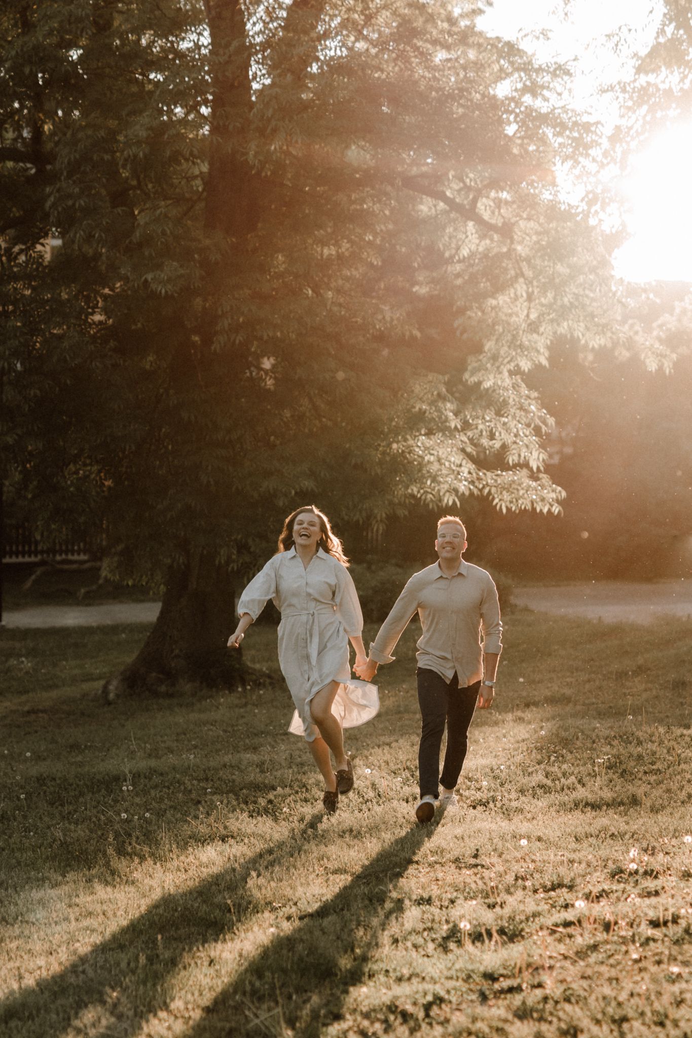 Joyful couple holding hands running through a park bathed in warm golden sunset light.