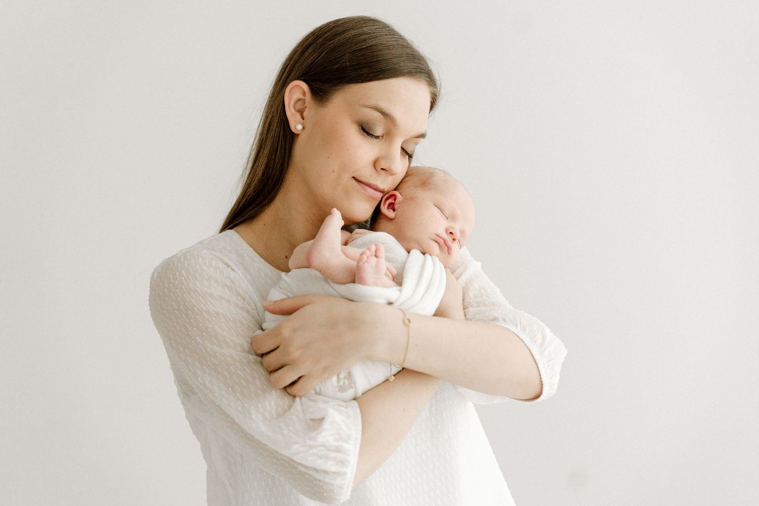 Young mother in white dress tenderly holding and cuddling her sleeping newborn baby.