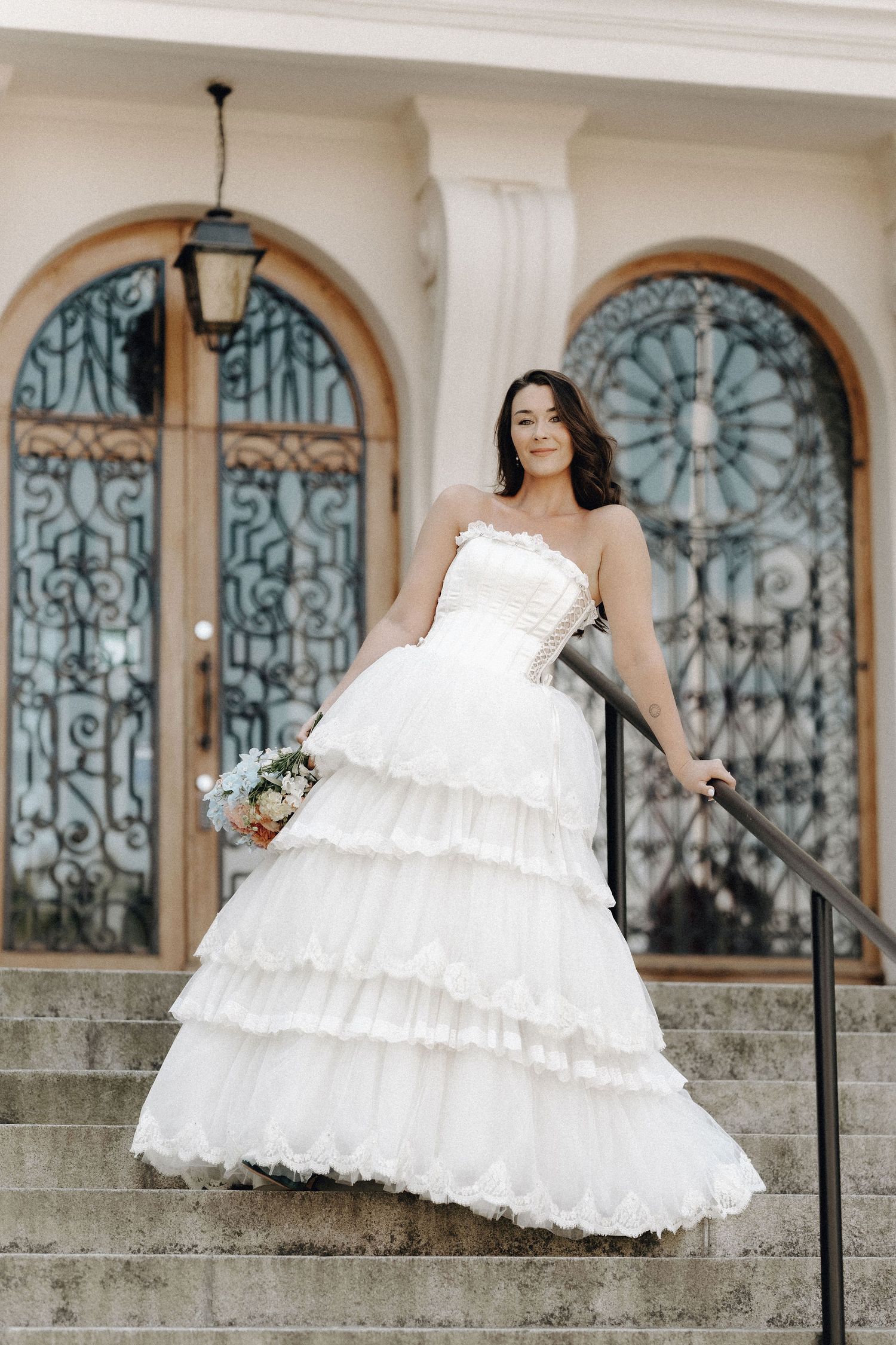 Elegant bride in tiered white strapless gown holding bouquet on steps of a grand ornate building.