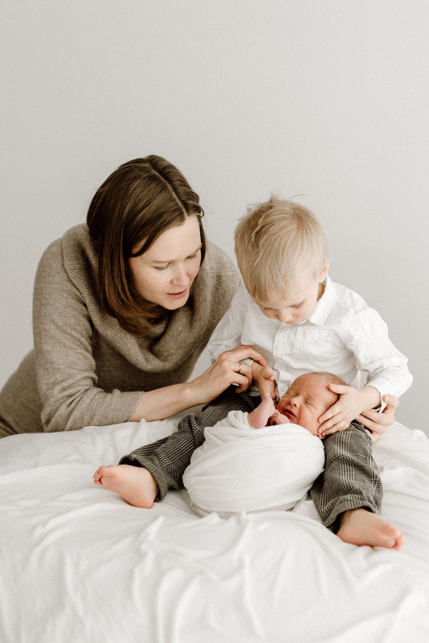 A mother and toddler boy sit on a white bed together admiring a swaddled newborn baby.