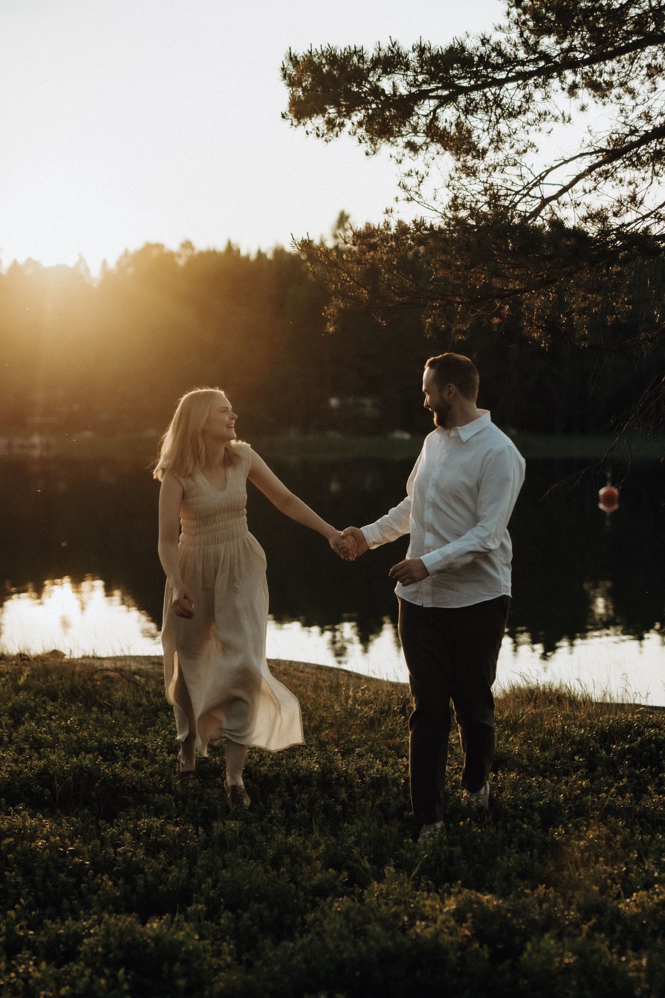 Couple holding hands walking by a sunlit lake at golden hour, woman in flowing dress, man in white shirt.