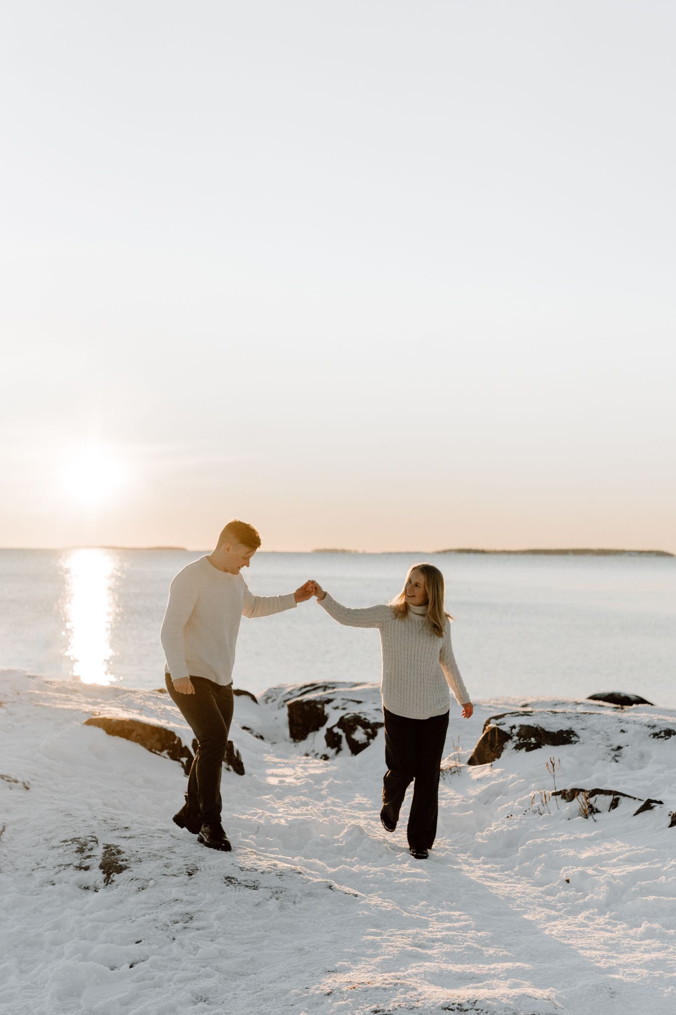 Romantic couple hold hands walking on a snowy beach with golden sunset reflecting on the water.