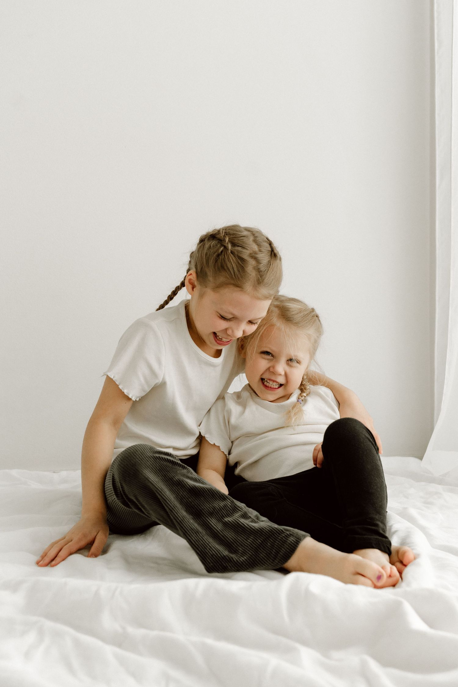 Two sisters with braids sitting on white bed, older girl hugging laughing younger sibling.