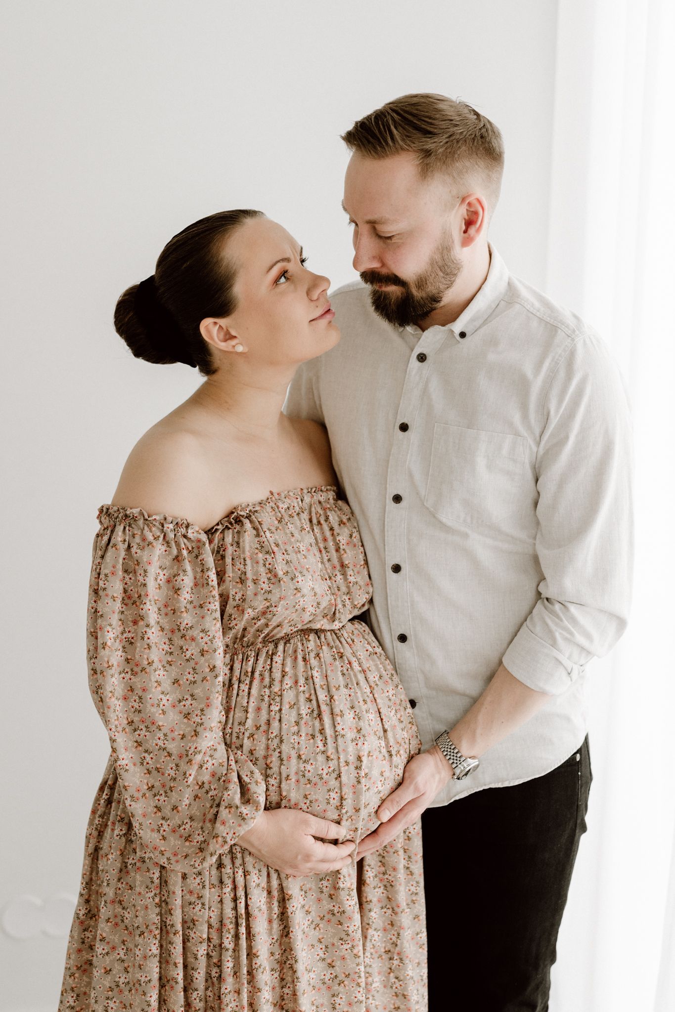 Pregnant couple tenderly embracing, woman in floral off-shoulder dress, man in white shirt, holding baby bump together.