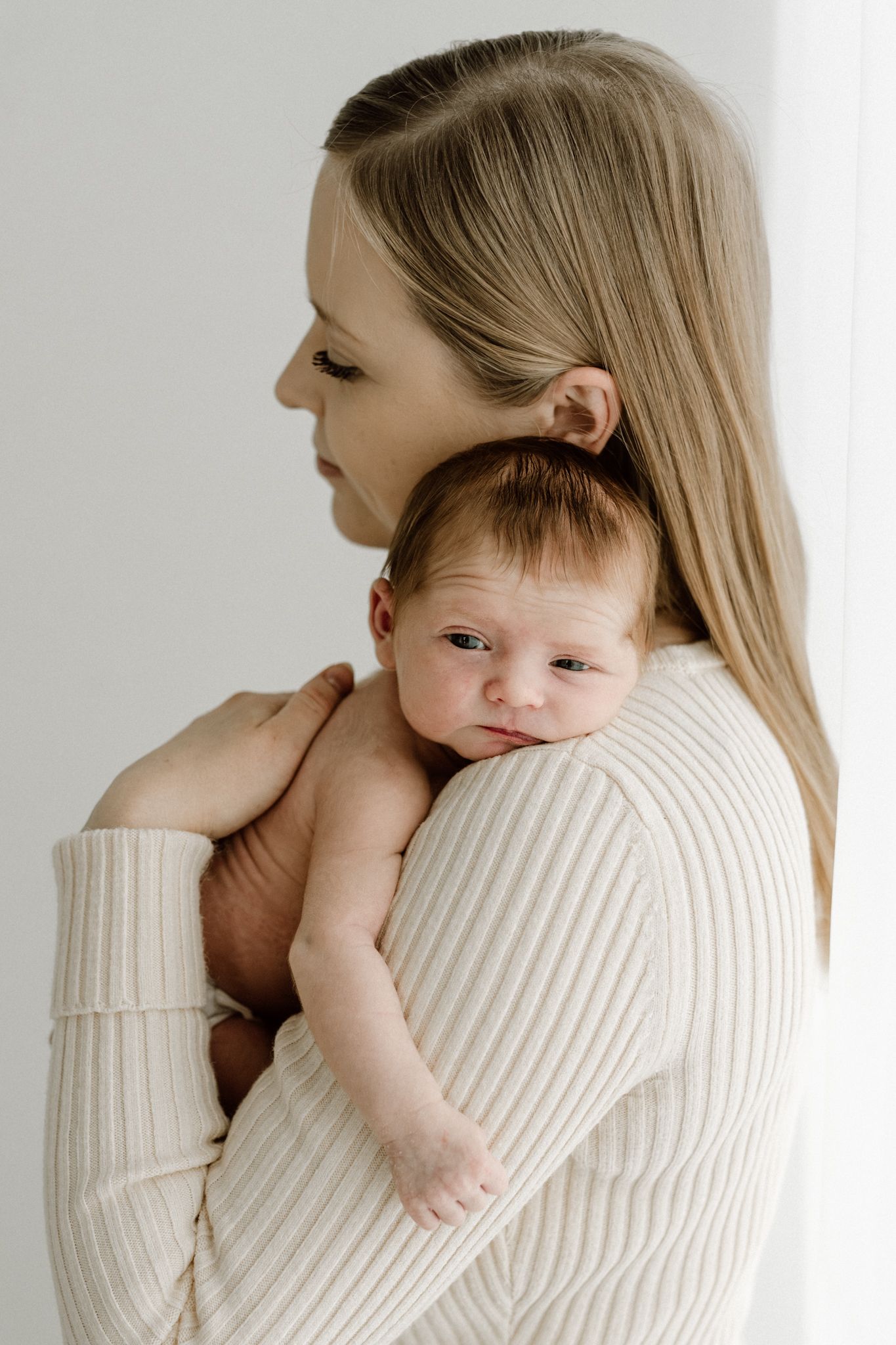 Mother in cream ribbed sweater holds alert newborn baby against her shoulder.