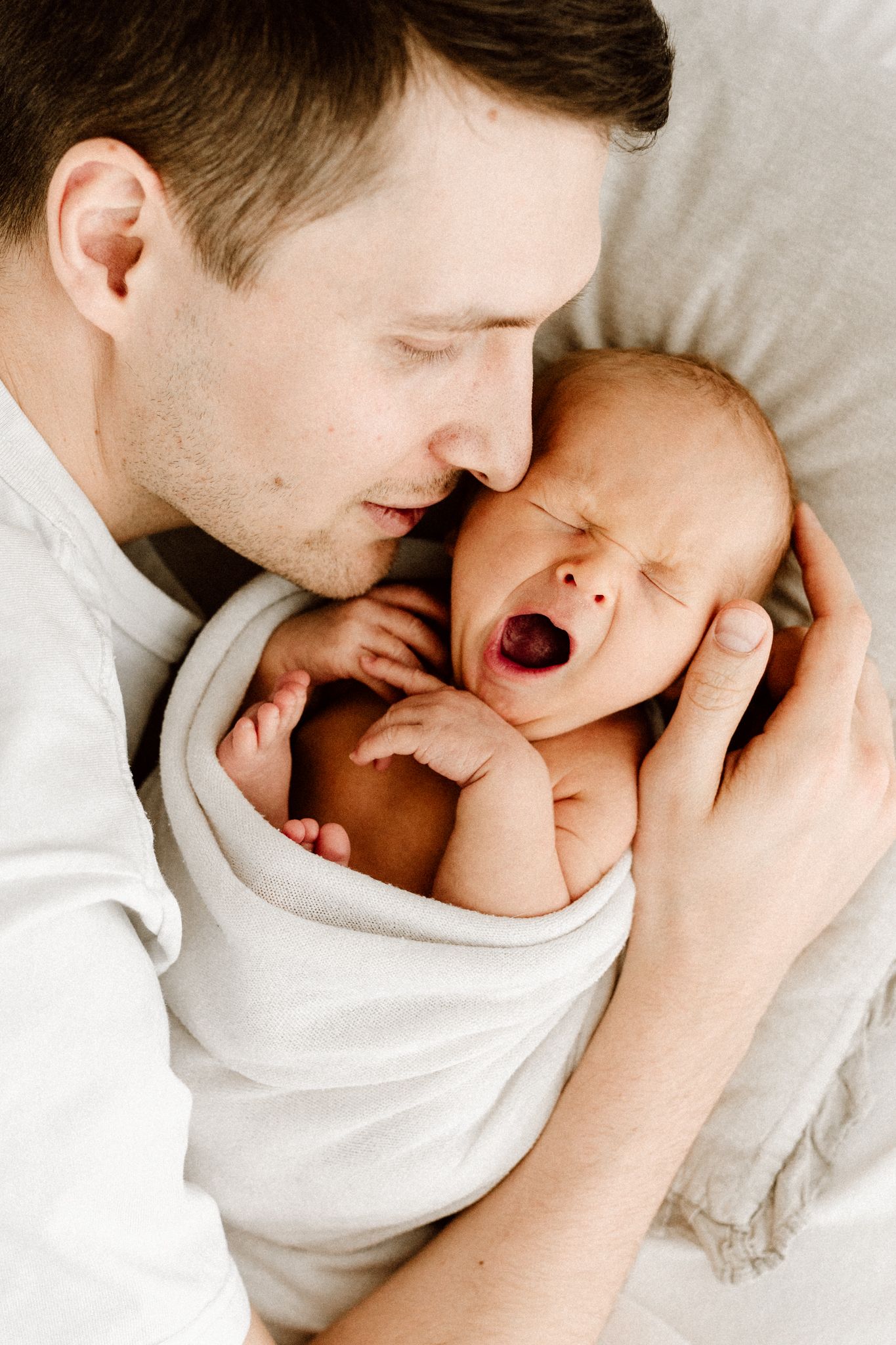 Father lying down holding his yawning swaddled newborn baby close to his face on white bedding.