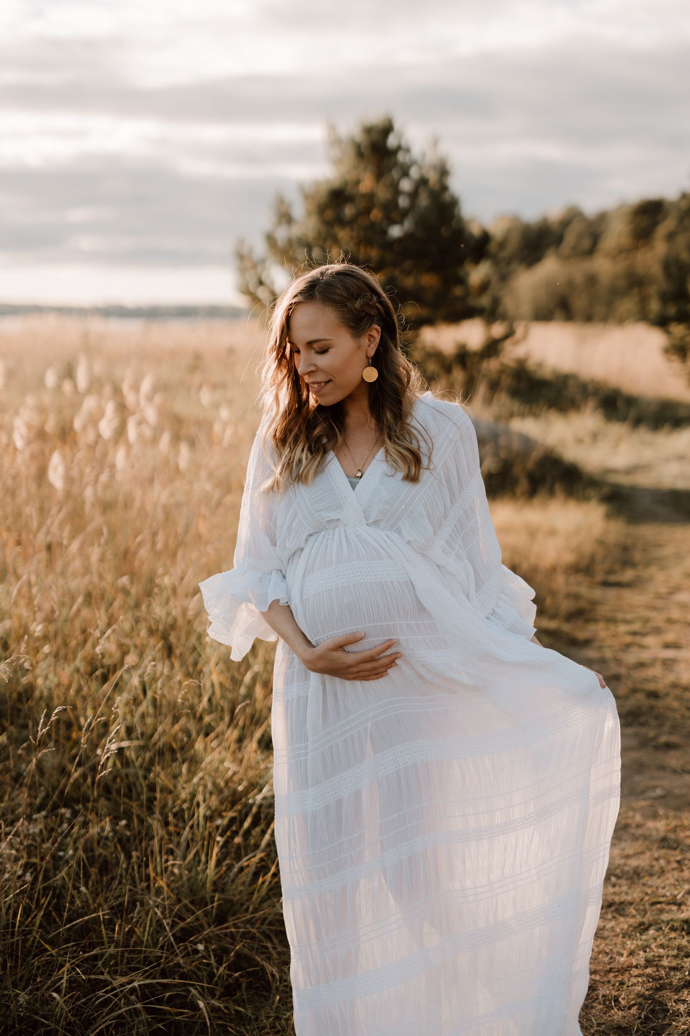 Pregnant woman in white dress looking down at baby bump in sunlit field during maternity session.