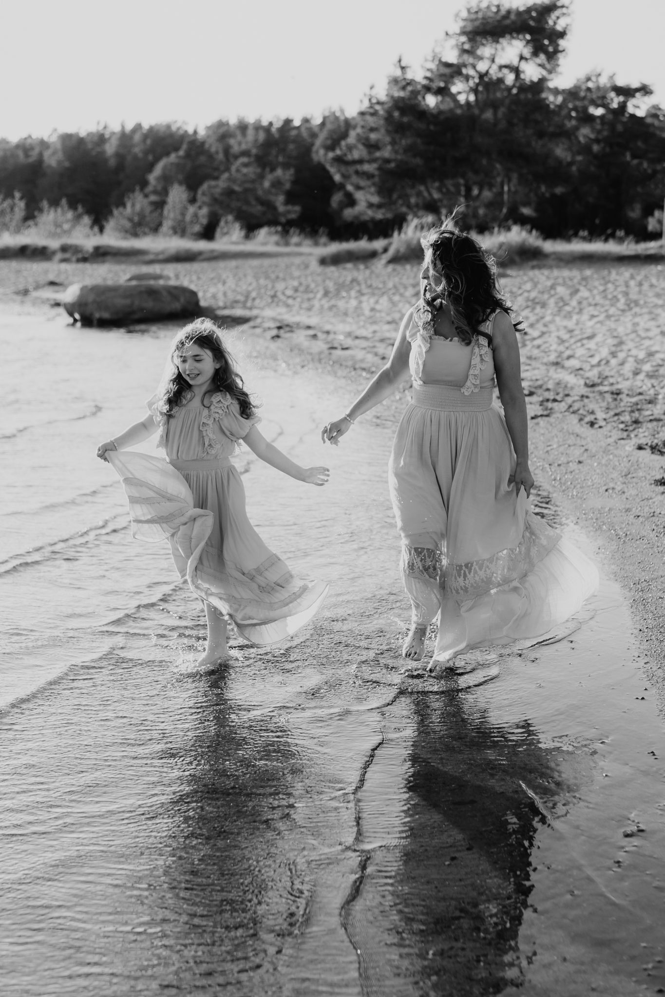 Black and white photo of a mother and daughter in flowing dresses running joyfully along the shoreline.
