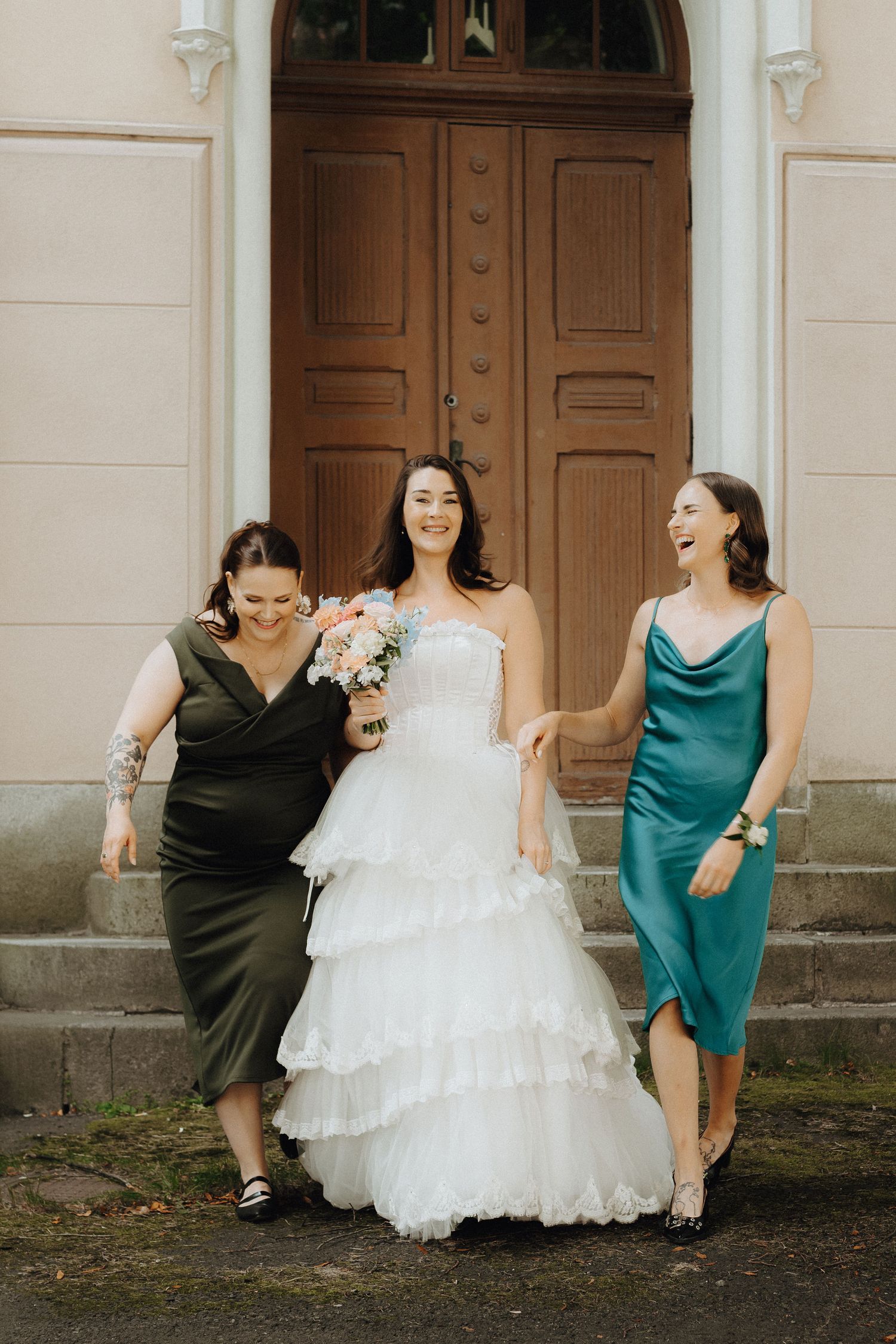 Joyful bride in ruffled white gown walks with bridesmaids in green and teal satin dresses outside a building.