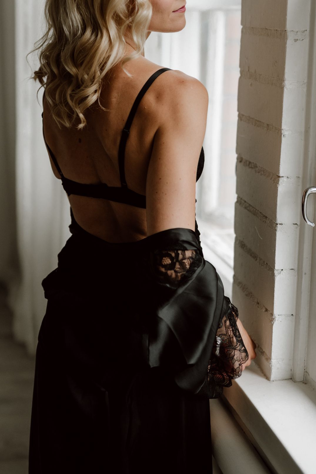 Woman in black lingerie and satin robe looking out bright window.