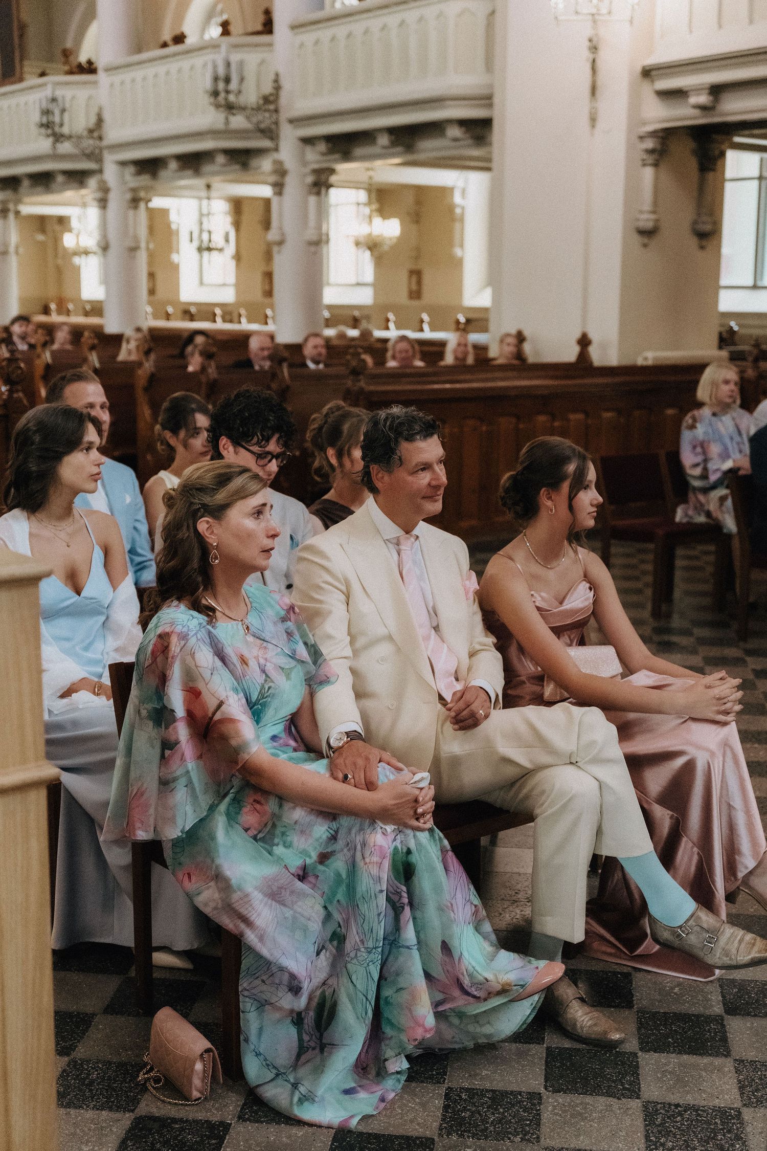 Wedding guests seated in church pews, woman in colorful floral dress in foreground smiling.