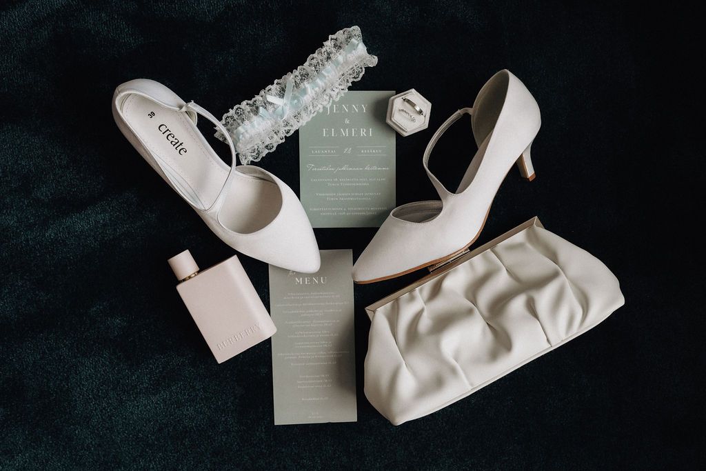 Close-up flat lay of wedding accessories featuring white heels, clutch, garter, and sage green invitations.