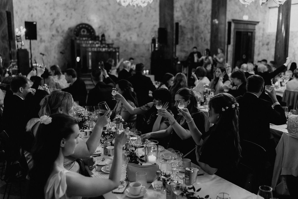 Black and white photo of wedding guests raising glasses in a toast during the reception dinner.