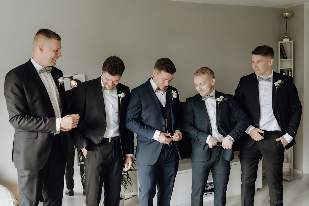 Five groomsmen in formal suits examine wedding accessories during pre-ceremony preparations.