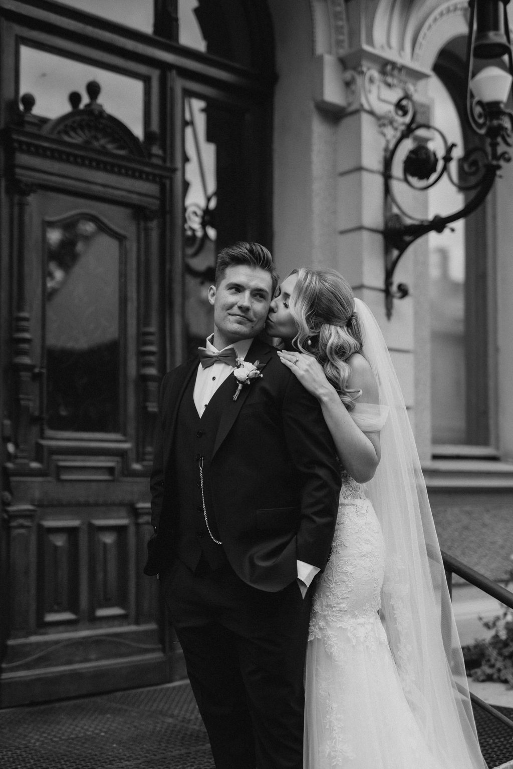 Black and white portrait of groom in tuxedo standing before ornate door while bride kisses his cheek.
