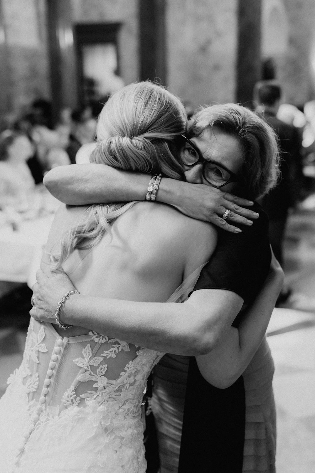 Black and white close-up of bride sharing heartfelt hug with glasses-wearing wedding guest.